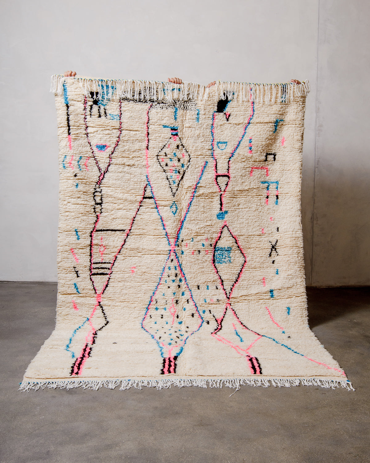 Berberlin Azilal rug made of fluffy cream wool and colourful design, held up against a white wall and cement floor.